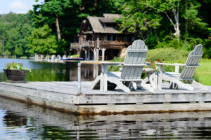 Two Adirondack chairs on wooden deck overlooking lake with cabin and boat dock in background.